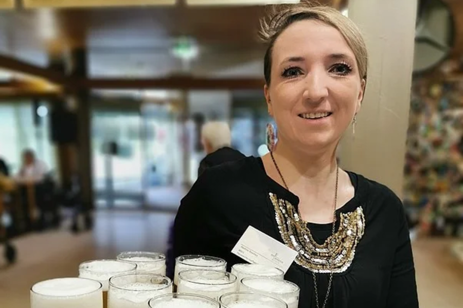 Kellnerin trägt schwarze Bluse und grüne Schürze, hält Tablett mit mehreren Gläsern Bier mit Schaum.