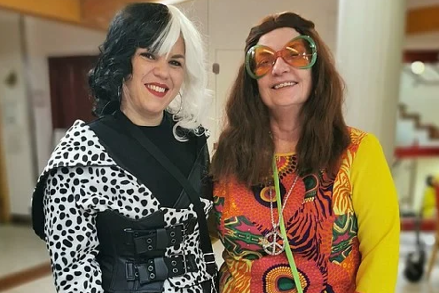 Zwei Frauen stehen nebeneinander, eine trägt ein schwarz-weiß gepunktetes Kleid mit schwarzem Gürtel und Handschuhen, die andere ein bunt gemustertes Kleid mit gelben Ärmeln und schwarzen Sneakers.