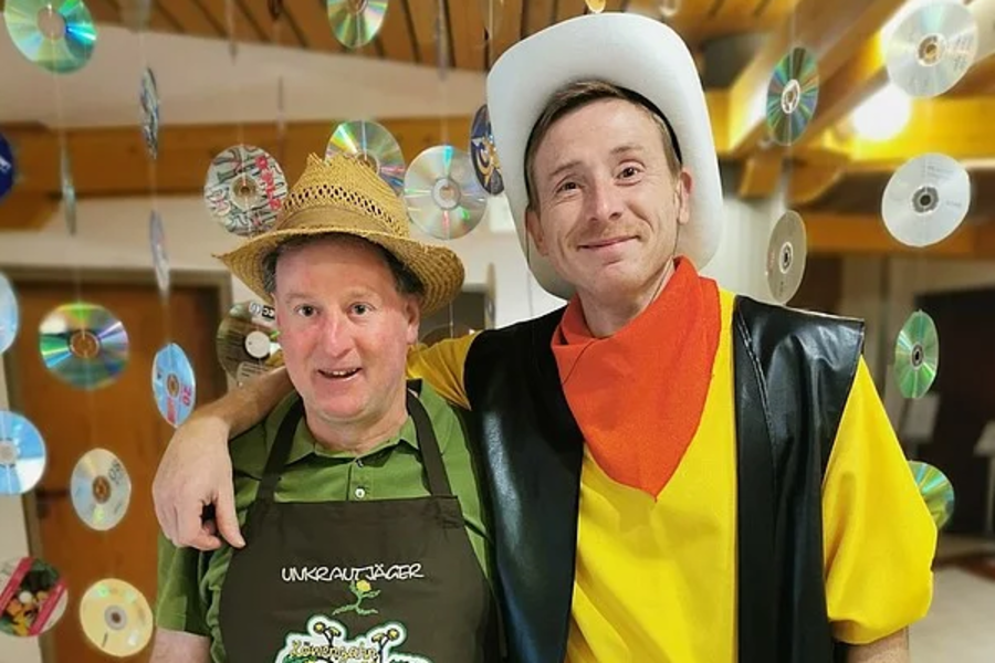 Zwei Personen stehen nebeneinander, eine trägt Strohhut und dunkelgrüne Schürze mit Aufdruck, die andere gelbes T-Shirt, schwarze Weste, orange Halstuch und weißen Cowboyhut.