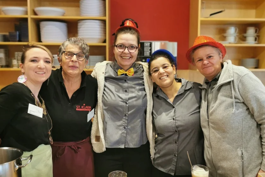 Fünf Personen in Arbeitskleidung und Schürzen stehen vor einem Regal mit Geschirr in einer Küche oder einem Restaurant.