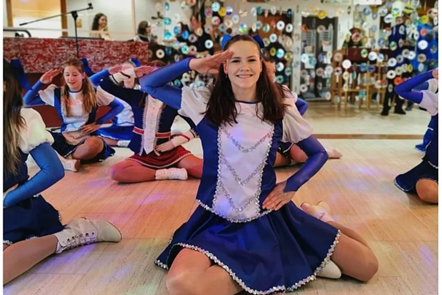 Junge Tänzerinnen in blauen und weißen Kostümen mit langen Ärmeln und kurzen Röcken bei einer Tanzaufführung in einem Raum mit Holzboden und Spiegelwand.
