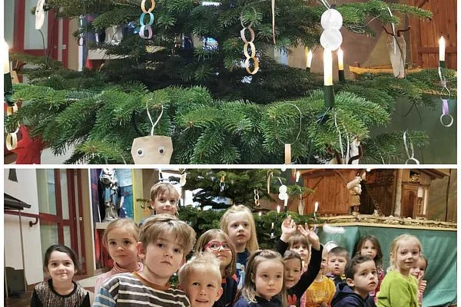 Weihnachtsbaum mit selbstgebasteltem Schmuck aus Papier und Holz, davor sitzende Kinder in bunter Kleidung in einem Raum mit Holzverkleidung.