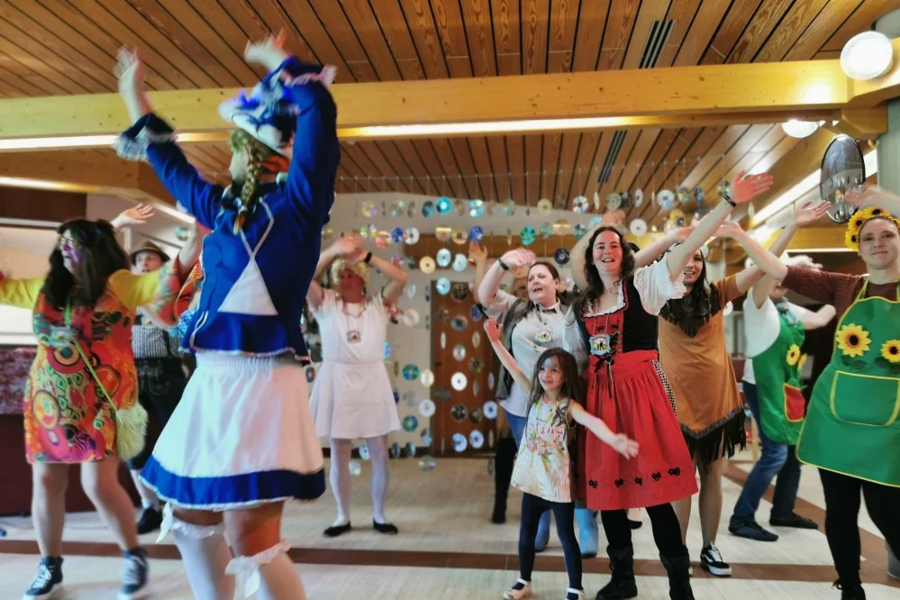 Personen in bunten Kostümen tanzen in einem Saal mit Holzboden und Holzwand, darunter ein blau-weißes Uniformkostüm und ein rotes Kleid mit schwarzen Punkten.