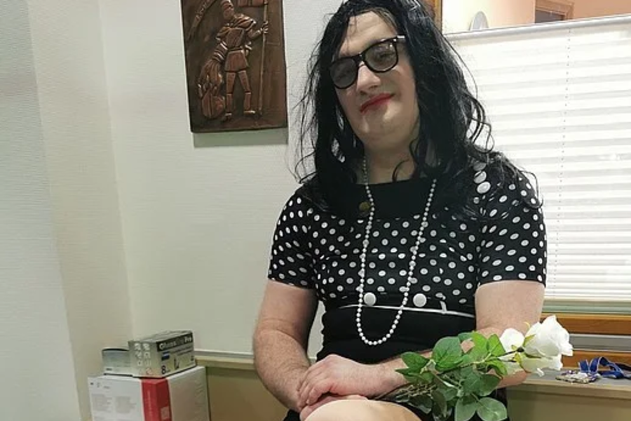 Frau in schwarzem Kleid mit weißen Punkten und Perlenkette sitzt auf Schreibtisch, hält weißen Rosenstrauß, trägt schwarze Schuhe mit Riemchen.
