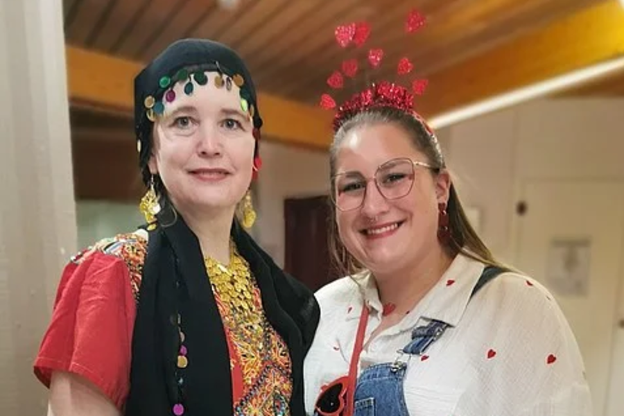 Zwei Frauen stehen nebeneinander, eine trägt ein rotes Kleid mit buntem Muster und schwarzen Schal, die andere blaue Latzhose, weiße Bluse mit roten Punkten und rotes Herzförmiges Täschchen.