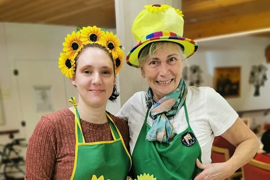 Zwei Personen tragen grüne Schürzen mit Sonnenblumenmotiven, eine mit gelbem Hut und bunter Borte, die andere mit Haarreif aus Sonnenblumen.