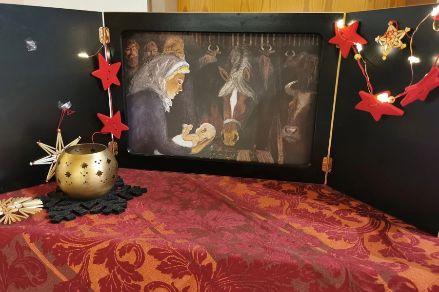 Krippe mit Figuren von Maria, Jesuskind und Tieren, umrahmt von roten Sternenlichtern und goldener Kerzenhalter auf rot gemustertem Tuch.