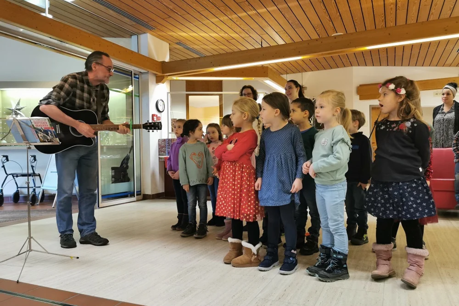 Mann spielt Gitarre vor einer Gruppe Kinder, die in einem Raum mit Holzdecke stehen und zuhören.