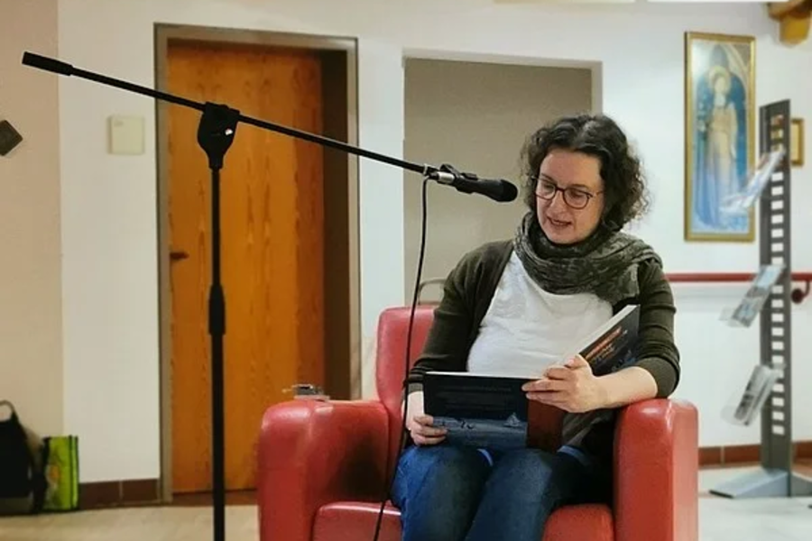 Person mit dunklem, lockigem Haar sitzt in rotem Sessel, trägt weiße Schuhe, blaue Jeans und weißes T-Shirt mit dunklem Cardigan, hält Tablet, davor Mikrofon auf Stativ.