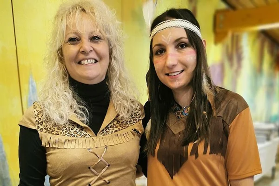 Zwei Frauen in braunen Kleidern mit Fransen, eine mit schwarzem Langarmshirt darunter, stehen nebeneinander in einem Raum mit gelb-grüner Wand.