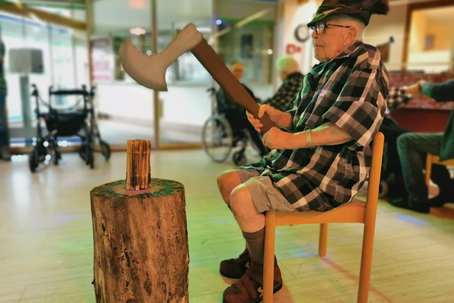 Kind sitzt auf Stuhl und hält Axt über Holzstamm mit Holzscheiten in Innenraum mit Rollstuhl und weiteren Personen im Hintergrund.