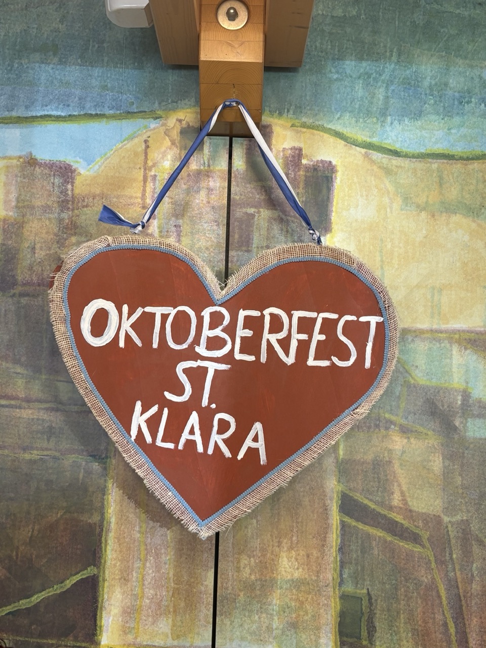 Braunes Lebkuchenherz mit weißer Schrift 'OKTOBERFEST ST. KLARA' und blau-weißem Band, hängend vor bemalter Wand.